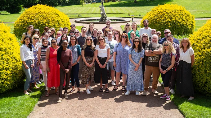 Group of about 45 smiling people standing together in the sunshine in formal gardens - composed of trainees and trainers on the Catalyse CAT Practitioner training course, double cohort 2022 - 2024