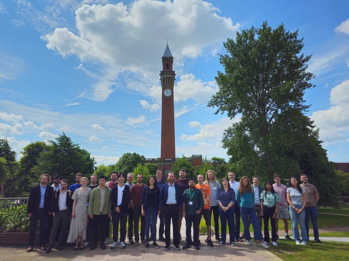 A group of about 30 people stand outside, with the Birmingham University clock tower behind them. The clock tower is tall and narrow and made of red brick. There are trees and bushes behind the group, and the sky is blue with white fluffy clouds. Most in the group are dressed for the warm day!