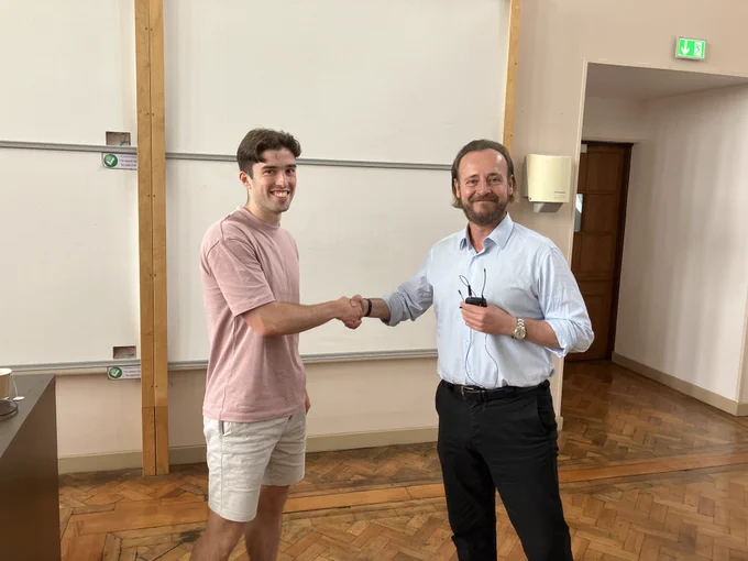 Poster prize winner Ollie stands at the front of a lecture theatre shaking hands with consortium leader Oliver!