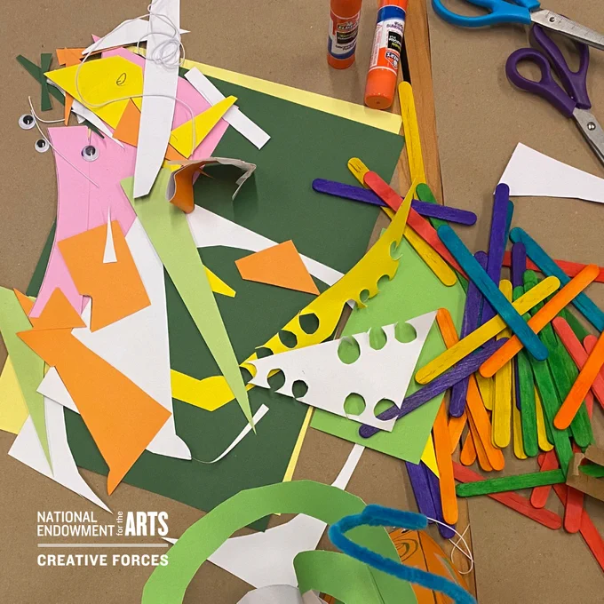 A view of a tabletop covered in brown kraft paper with art supplies on top, including pieces of paper in different colors cut in different shapes, googly eyes for art projects, multi-colored popsicle sticks, glue sticks, and scissors.