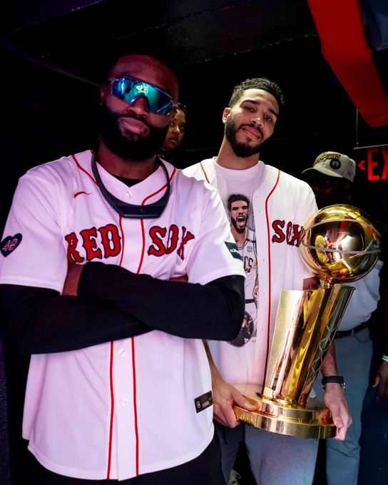 Jayson Tatum and Jaylen Brown pose with the Larry O'Brien Trophy in the batting tunnels at Fenway Park, wearing Red Sox jerseys.