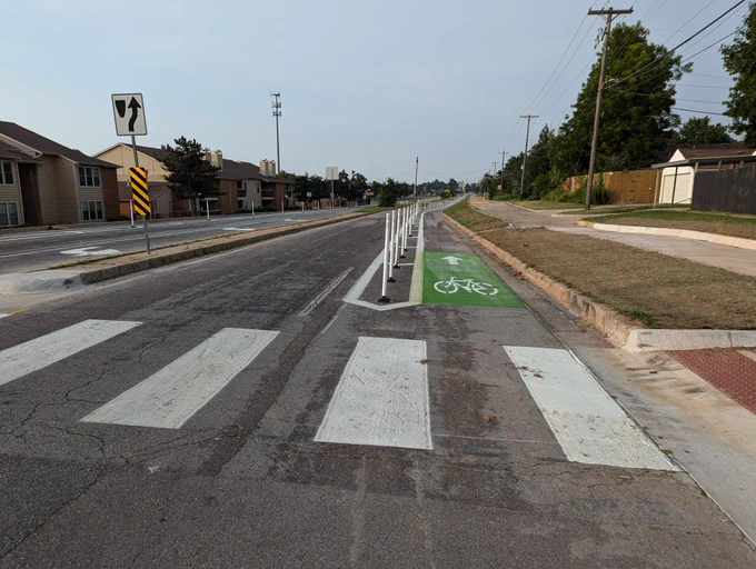 On the other side of the street, a similar photo of fresh crosswalk, ADA-,compliant curb cut for sidewalk access, flex-post protected bike lane next to a car traffic lane.