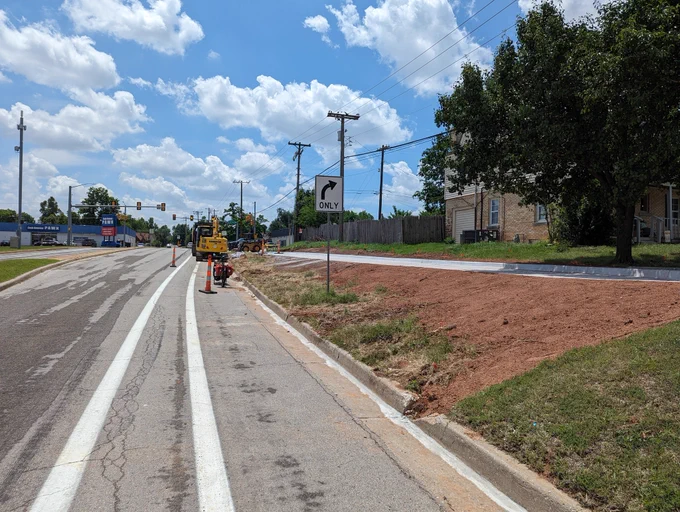 View from the bike lane with cones and a construction vehicle ahead. The grass to the side is sloped upward, and the white of fresh-paved sidewalk can be seen gently grading down from the higher neighborhood to meet up with the bike lane further ahead.