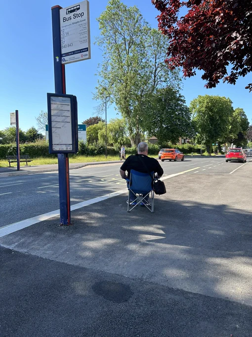 Man sitting in a foldable camping chair at a bus stop with no shelter or seating.
