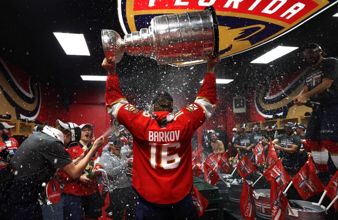 Photo of Panthers captain Sasha Barkov from behind holding the Stanley Cup above his head.

He is in the locker room with his teammates spraying champagne all around him.