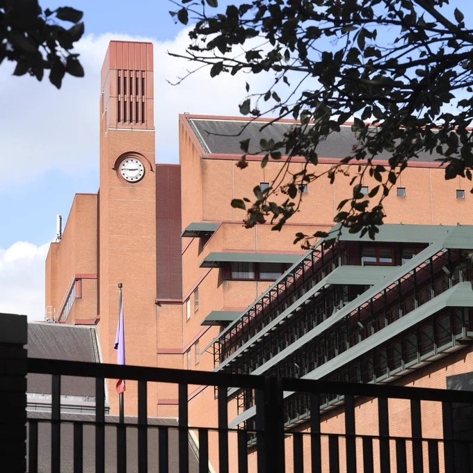 A photograph of the exterior of the British Library at St Pancras with the clock tower and adjacent building in view, framed by tree branches and the top of a silhouetted fence.