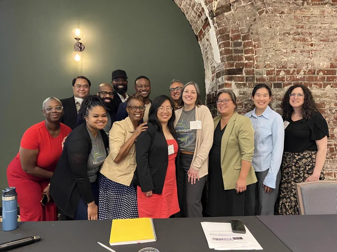 Group picture with 13 people smiling at a conference table in front of a brick wall.
