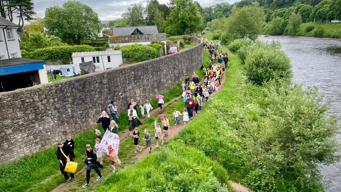 A straggly line of children and adults in costume makes its way along a path between the river bank and an old stone wall in Usk, lead by flags and a samba band. 