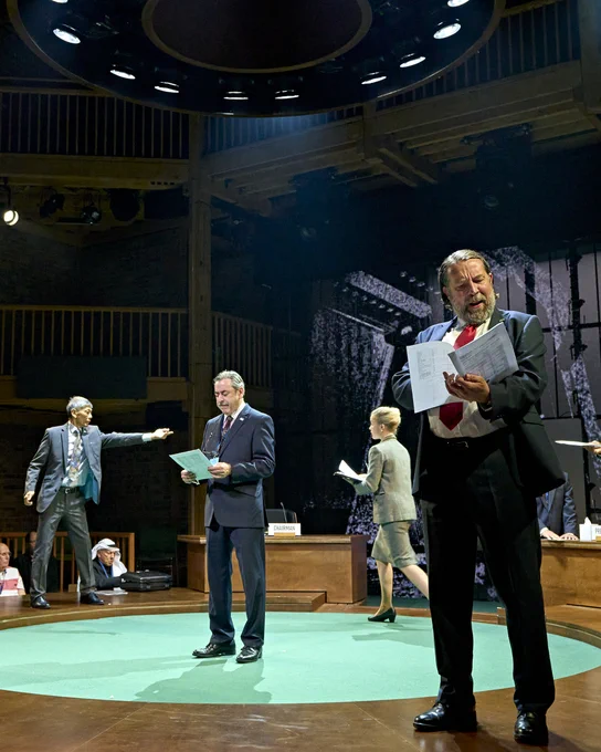 People in suits stand on a circular wooden table reading paper reports, while other people distribute the reports among delegates.