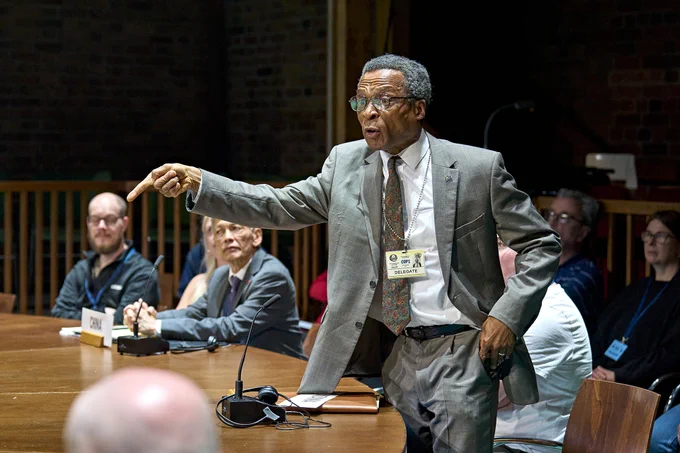 Jude Akuwudike wears a grey suit and lanyard and stands around a conference table with right arm raised, index finger pointing forwards. 