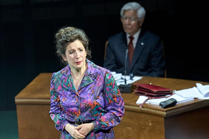 Jenna Augen stands wearing a flowery blue blouse with hands clasped. Behind, Togo Igawa sits at a desk littered with papers and folders.
