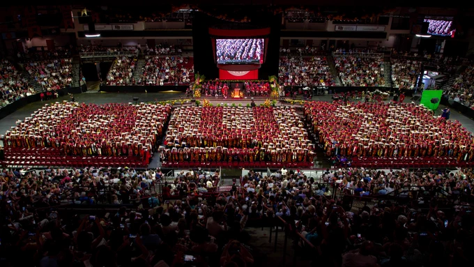 The Class of 2024 stands facing the audience in Magness Arena during Commencement. The graduates are holding red and white pennants that read "DU '24" from an aerial view.