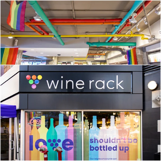 This image is a collage of two separate photos related to a wine store named "Wine Rack." The top photo depicts the interior of the store with multiple colorful flags stretched across the ceiling, creating a vibrant and celebratory atmosphere. The lower photo shows the exterior of the store with a "Wine Rack" sign displayed prominently above the window. The window features a display with large, colorful illustrations of wine bottles and glasses, and text that reads "shouldn't be bottled up" and "Your neighbourhood pride spot." The open sign is visible in the window, signaling that the store is open for business.
