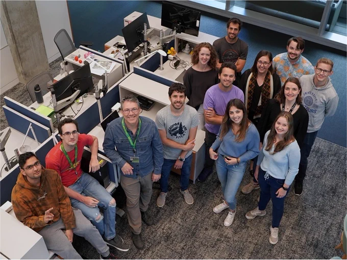 Group of thirteen colleagues smiling for a photo in a modern office environment.