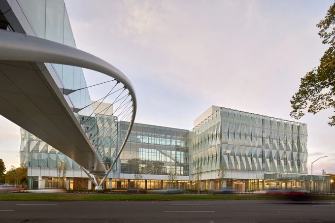 Modern architectural building featuring a large glass facade with a unique, circular steel structure in the foreground that appears to support a bridge or walkway. The building is surrounded by a well-manicured lawn and a roadway with slight motion blur indicating passing traffic. Clear sky visible above during what appears to be either dawn or dusk.