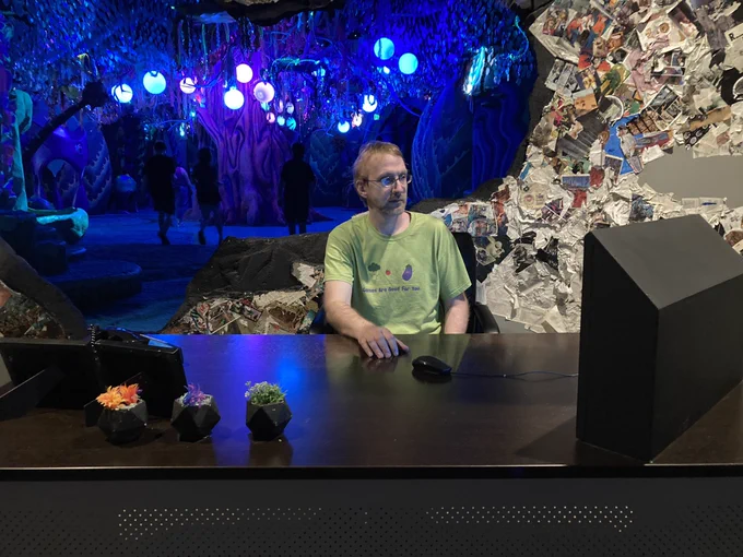 Shawn works at a desk, with a broken wall behind him, leading to an inside grotto of hanging lights and people walking around