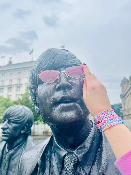 A hand placing pink heart-shaped sun glasses on a statue of John Lennon.
