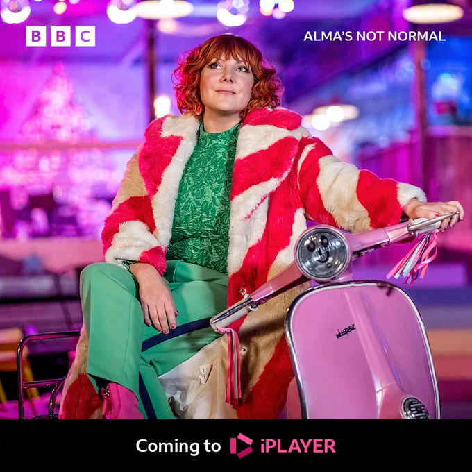 Alma sits on a pink scooter, wearing a green top and trousers, pink books and a furry striped pink and white coat.