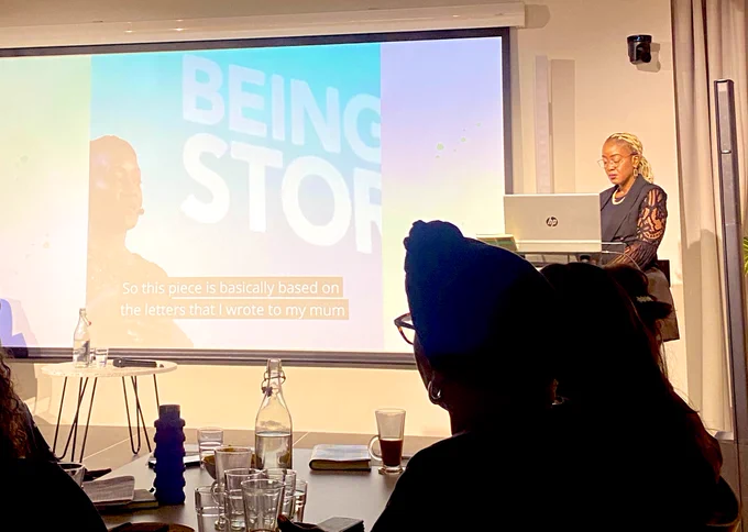 Photo of a woman on stage she is in her 30s black with long blond braids. There is a screen showing a video of her giving public talks
