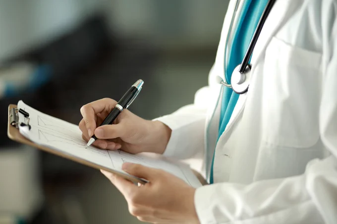 A healthcare professional wearing a white lab coat is writing on a medical chart attached to a clipboard.