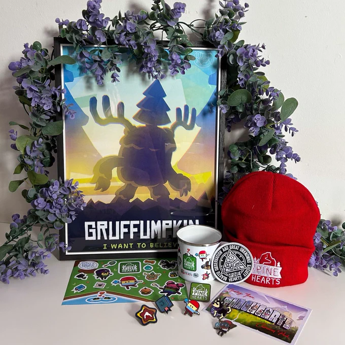 Gruffumpkin poster in a frame leaning up against a white wall with eucalyptus and purple flowers draped over it. Below the frame are a collection of items including: A red Pine Hearts embroidered beanie, a white enamel camping mug featuring characters from Pine Hearts, a sticker sheet, a postcard, a patch, and 6 enamel pins.