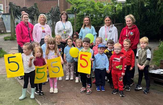 A group of young children and adult teachers hold up colourful pieces of card on which are written the number five.