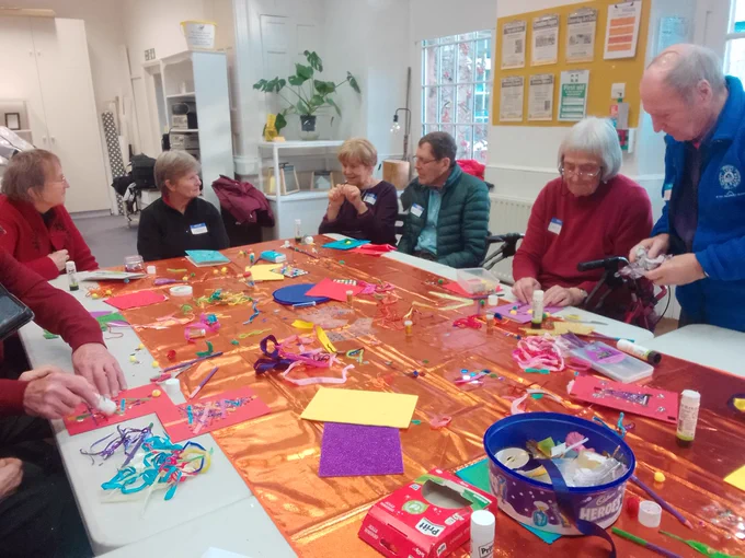 A group of older people sit around a busy table covered with brightly coloured art materials. Some of them are chatting, others are cutting and sticking