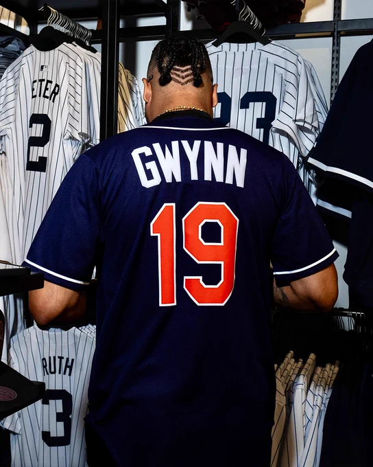 Luis Arraez puts on a retro Padres Tony Gwynn jersey. He has his back turned to the camera as he shows the #19 and lettering.
