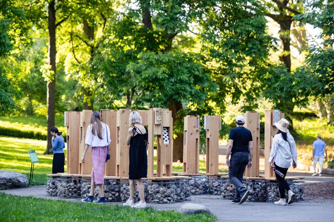 Six or so people walking around an art installation made of tall pieces of wood and metal cube bases filled with stone. The installation can be found in a forested outdoor space in a botanic garden.