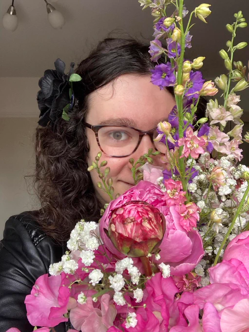 A picture of me, smiling, half-hiding behind a gigantic bunch of flowers in pastel shades (including sweet peas and roses). I am wearing a leather jacket (only the sleeve is visible) and a black flower in my long curly brunette hair.