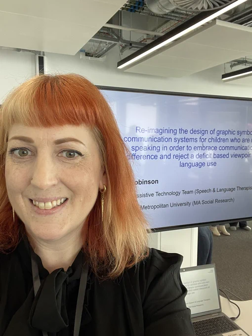 Helen is stood taking a selfie, smiling. She is wearing a black blouse and has shoulder length orange and blonde hair and is smiling. Behind her is a screen displaying her presentation.