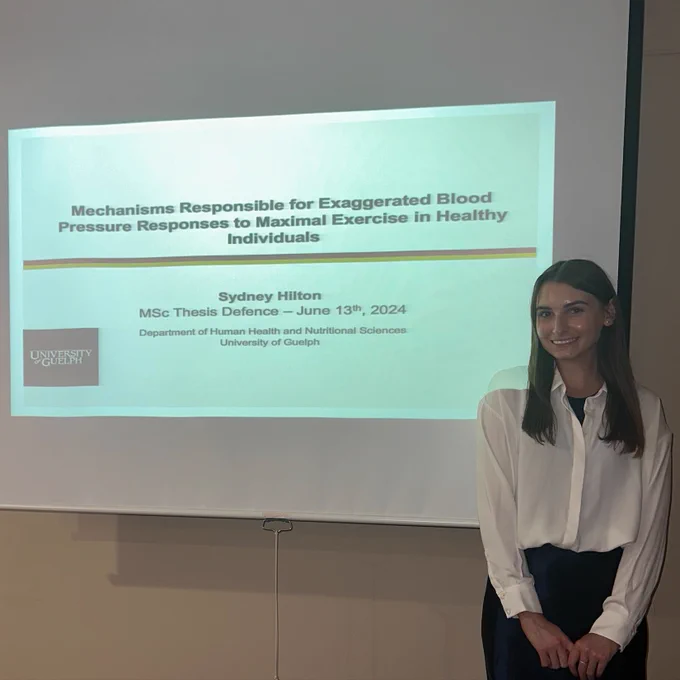 A photo of Sydney at her defence, in front of a projected title screen with the same title of her thesis that is listed in this posting.