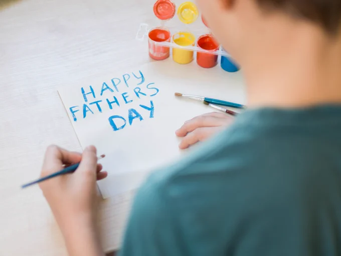 A student painted 'Happy Father's Day' in blue paint on a blank sheet of paper. Their paint palette is visible in the background.