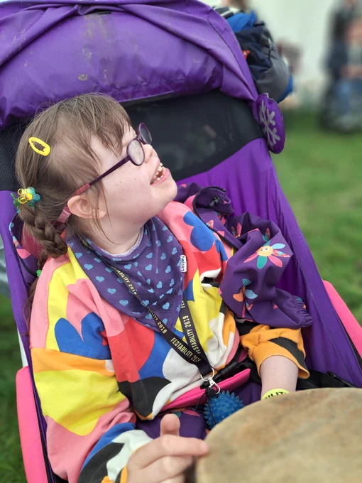 Disabled girl wearing purple glasses and bright clothes sits in a purple buggy and playsa drum. She's smiling and looking up to her left.