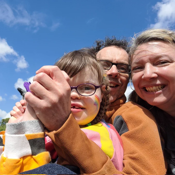 A mum, dad and their disabled daughter sit in a field, blue sky with white clouds overhead and smile into a selfie.