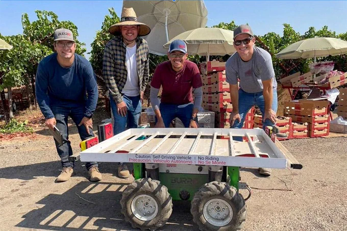 Four people stand behind a small, self-driving vehicle labeled in a vineyard setting. They are smiling, and the vineyard stretches out behind them under a sunny sky.