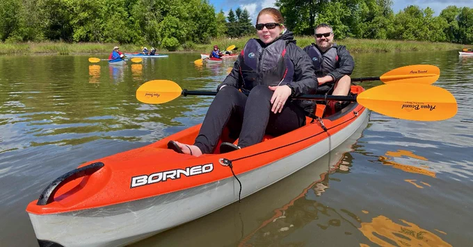 orange tandem kayak reads borneo, athlete in front and volunteer in back with yellow paddles posed in their laps, smiling at the camera both wearing black sunglasses and black leggings with black and red mti life jackets in the foreground, in the background clue skies with clouds and big beefy green trees