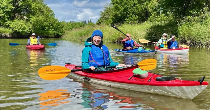 volunteer in a red kayak smiles at the camera, tandem kayaks and single kayaks in the background, cloudy blue skies on muddy bay water