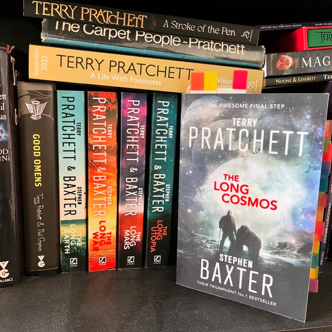 A photo of the book The Long Cosmos on a bookshelf. It’s on the right, cover facing us, next to the first four in the Long Earth series, spines out. Other Terry Pratchett books are also visible, including Good Omens, A Stroke of the Pen and the original edition of The Carpet People.