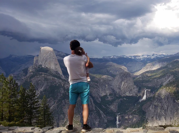 A father holds his young daughter close to him while he takes in a gorgeous view of Yosemite National Park’s soaring granite cliffs with waterfalls cascading down into a green, pine covered valley. 