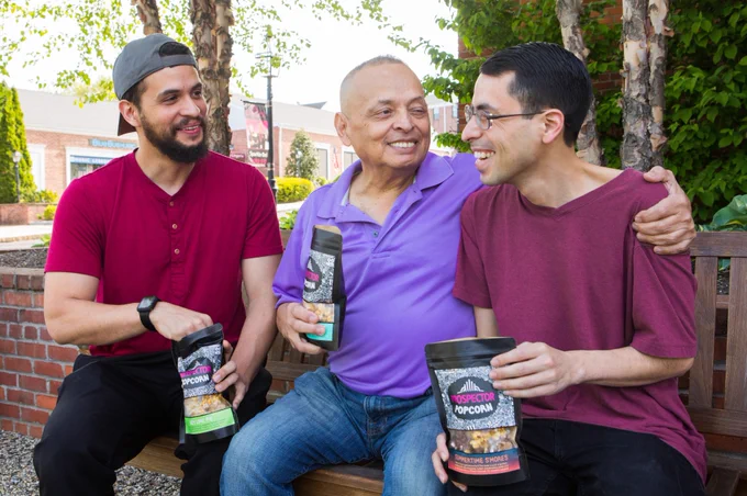 A father and his two sons smile while sharing three bags of gourmet popcorn together.