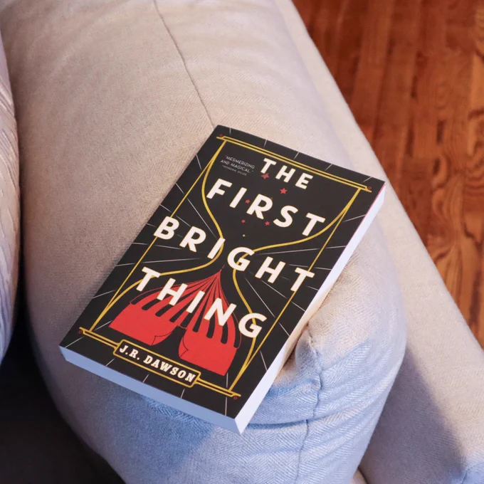 The First Bright Thing by JR Dawson is laying on a grey couch 