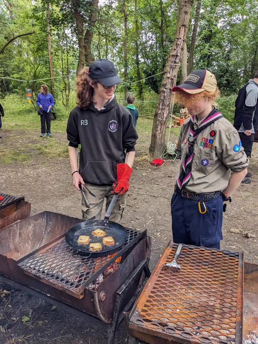Two young leaders stand by an altar fire (half a metal barrel), topped with a grill. One is holding tongs and a frying pan, which is on the grill and has camp donuts inside (jam sandwich covered in pancake batter)