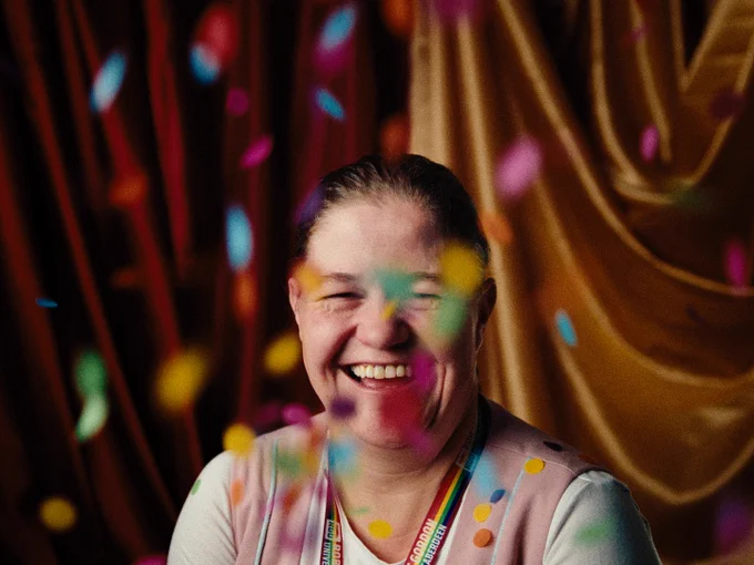 A women sits in front of colourful draped fabric and smiles as confetti falls all around her.