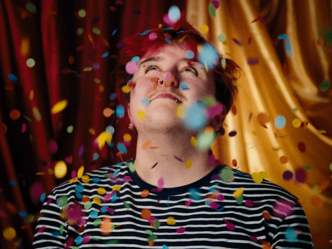 A man sits in front of colourful draped fabric and looks up as confetti falls all around him.