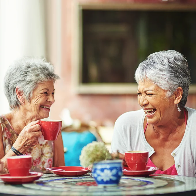 Two elderly women with short gray hair are sitting at a colorful, mosaic-tiled table, laughing while drinking from red mugs. The woman on the left is smiling warmly, while the woman on the right is laughing heartily.