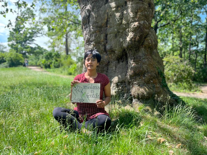 Neelakshi Joshi - the speaker - is sitting in a park on a meadow at the foot of a large tree. She is holding a cardboard sign in her hand: ‘Resistance is fertile’