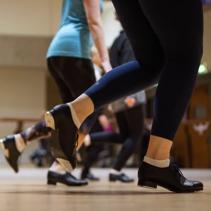 A close up photo of a group of people wearing exercise clothing and tap dancing shoes captured mid-movement. The camera is focused upon the lower half of their bodies with none of their faces visible. 