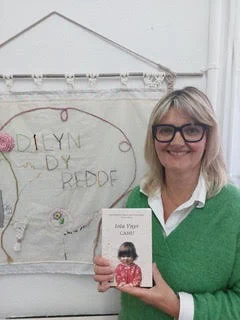 Picture of Iola Ynyr holding her book, Camu, in front of a banner that says 'Dilyn dy reddf' ('Follow your instinct') 