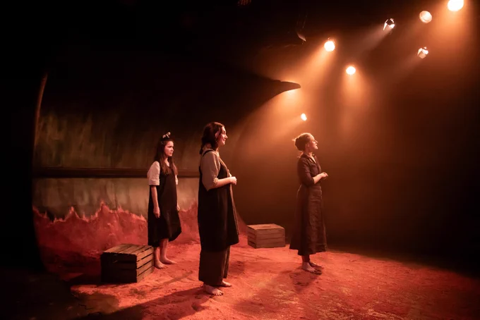 Three women stand side-on in a theatre, lit with warm orange-yellow light. The floor of the set is covered in red-orange dirt, and there are two wooden crates upstage. At the back, the floor blends seamlessly into a wall which curves up and over.